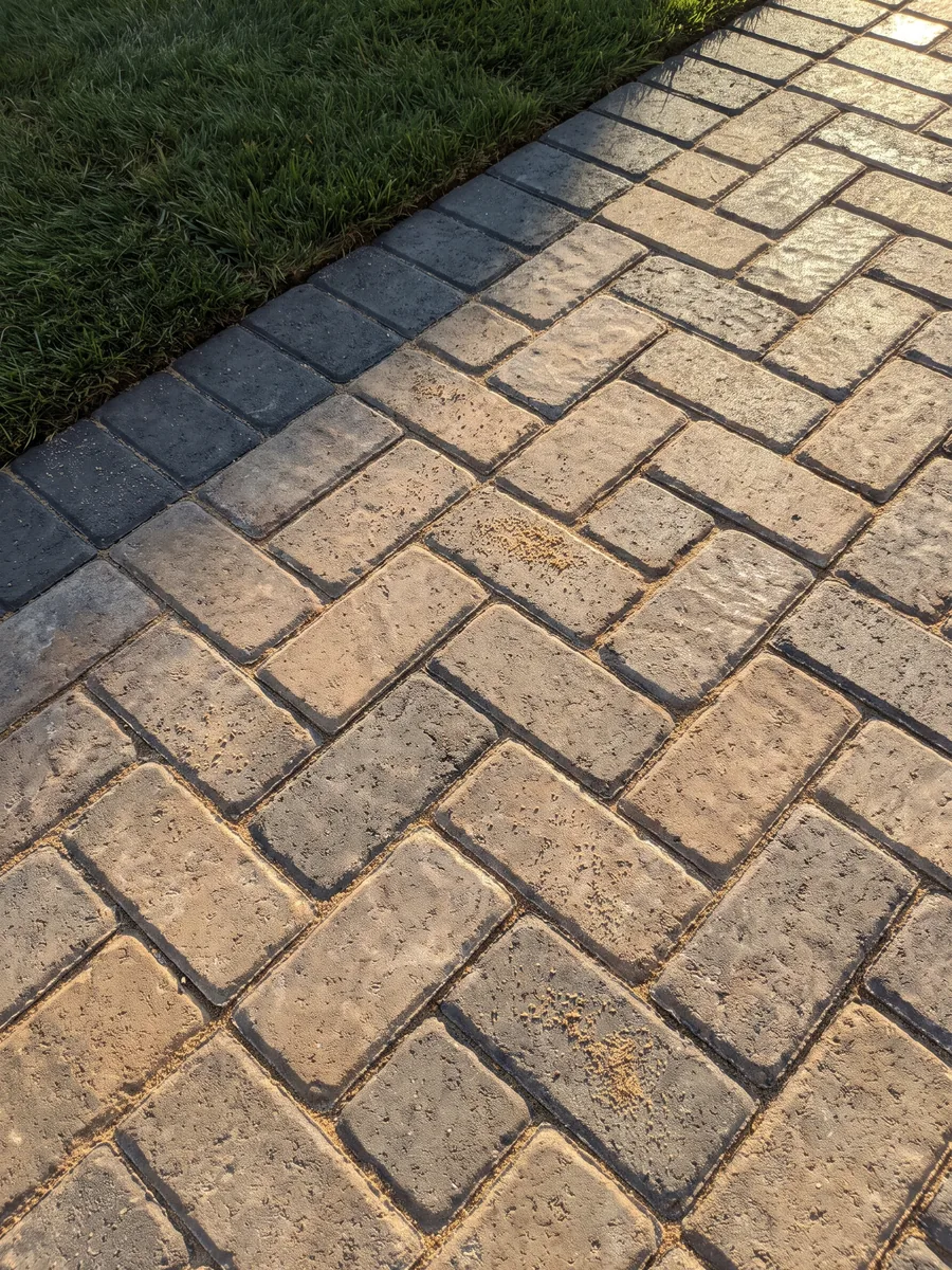 The corner of a finished herringbone paver patio with a charcoal border, joints swept, meeting new sod